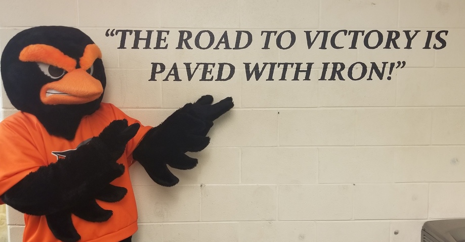 Oriole bird mascot points to sign in the CHS weight room that says, "The road to victory is paved with iron!"